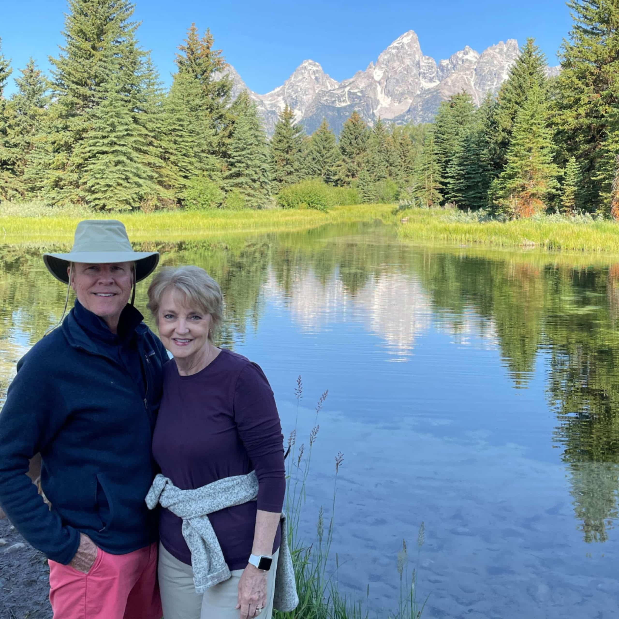 Two people standing in front of a pond with mountains in the background.