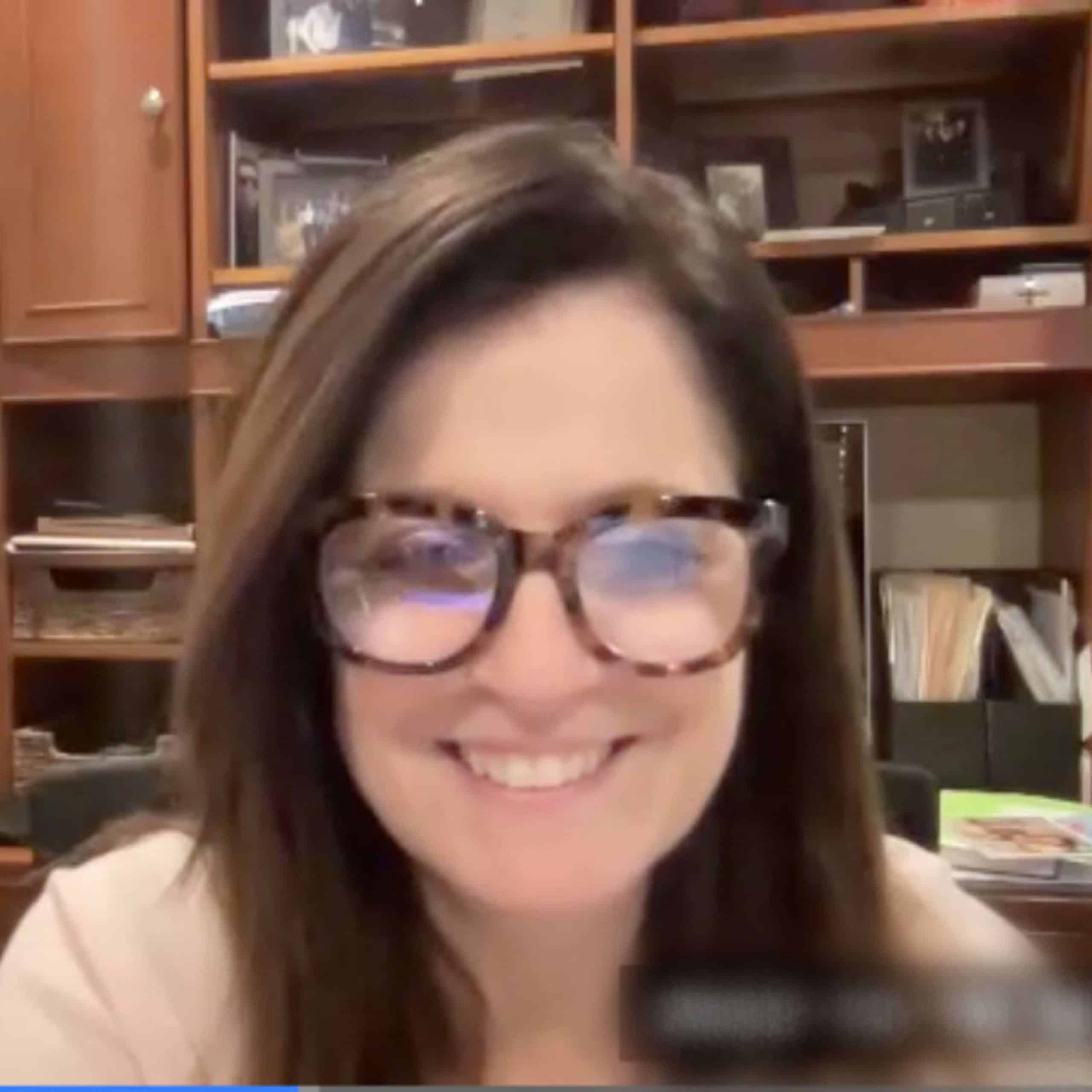 A woman wearing glasses is smiling in front of a desk.