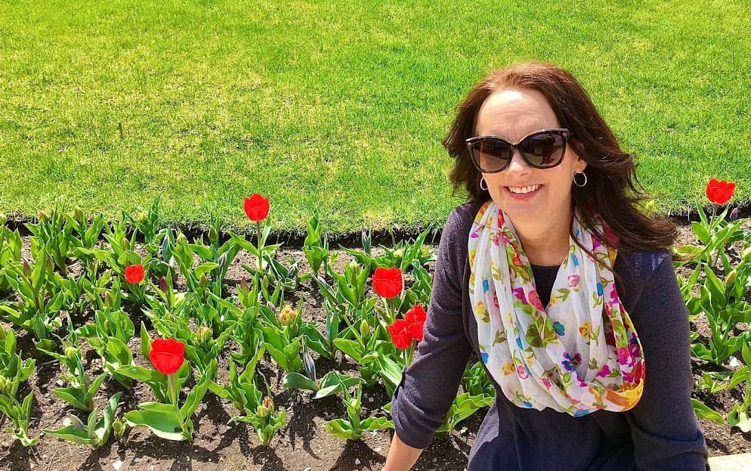 A woman wearing sunglasses and a scarf in front of a flower garden.